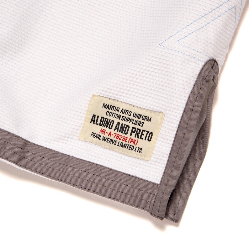 May include: A white martial arts uniform jacket with a textured weave, featuring a grey trim along the hem and a rectangular patch. The patch reads: MARTIAL ARTS UNIFORM COTTON SUPPLIERS ALBINO AND PRETO MIL-A-7823E (PK) PEARL WEAVE LIMITED LTD.