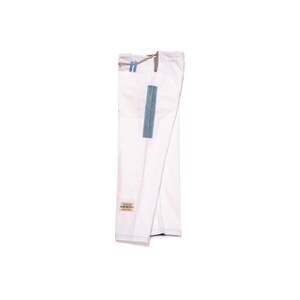 May include: A pair of white martial arts pants featuring a drawstring waist with light blue accents. A vertical blue rectangular patch is positioned on the side of the leg, and a small rectangular label with text is sewn near the bottom hem.