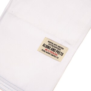May include: A white cotton martial arts uniform fabric featuring a rectangular cream-colored label with black and red text that reads: MARTIAL ARTS UNIFORM COTTON SUPPLIERS ALBINO AND PRETO MIL-A-7823E (PK) PEARL WEAVE LIMITED LTD. The fabric has visible stitching.