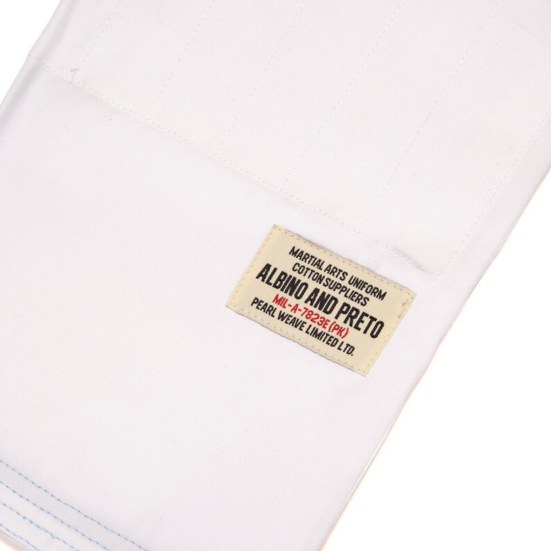 May include: A white cotton martial arts uniform fabric featuring a rectangular cream-colored label with black and red text that reads: MARTIAL ARTS UNIFORM COTTON SUPPLIERS ALBINO AND PRETO MIL-A-7823E (PK) PEARL WEAVE LIMITED LTD. The fabric has visible stitching.