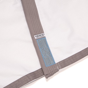 May include: A white martial arts uniform jacket featuring a textured weave fabric, grey trim along the lapel and hem, and a rectangular patch with the text ALBINO AND PRETO MARTIAL ARTS UNIFORM above a blue geometric pattern on the lapel.