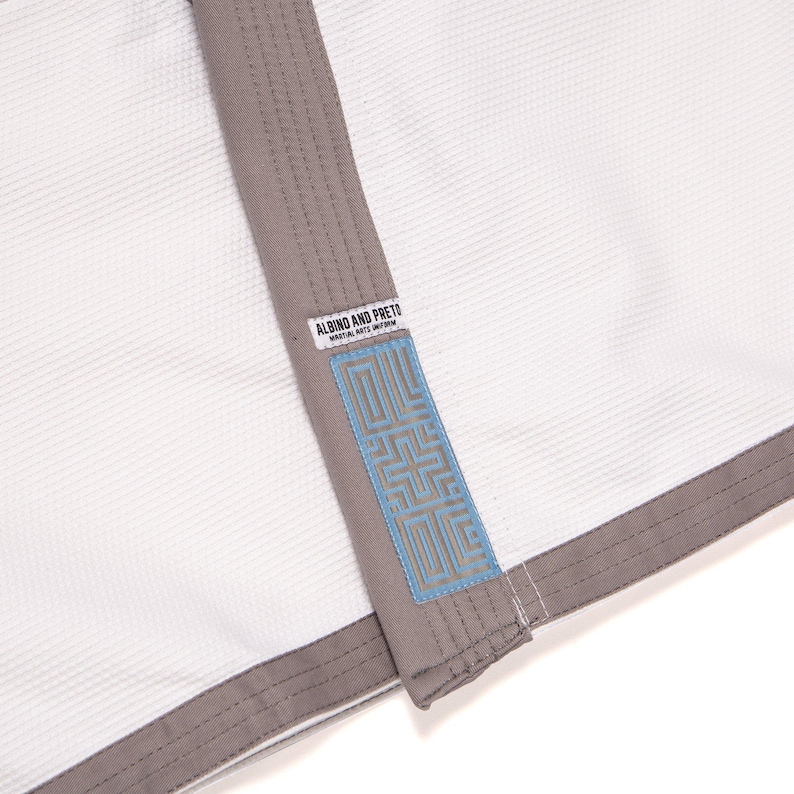 May include: A white martial arts uniform jacket featuring a textured weave fabric, grey trim along the lapel and hem, and a rectangular patch with the text ALBINO AND PRETO MARTIAL ARTS UNIFORM above a blue geometric pattern on the lapel.