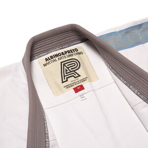 May include: A white martial arts uniform jacket featuring a grey collar and a cream-colored interior label with the text ALBINO&PRETO MARTIAL ARTS UNIFORMS, a logo, and a red A1 size tag. The fabric has a textured weave and blue trim details.
