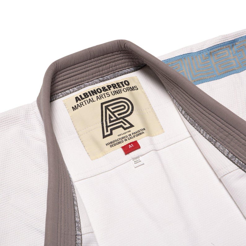 May include: A white martial arts uniform jacket featuring a grey collar and a cream-colored interior label with the text ALBINO&PRETO MARTIAL ARTS UNIFORMS, a logo, and a red A1 size tag. The fabric has a textured weave and blue trim details.