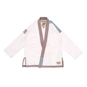 May include: A white martial arts gi jacket featuring a grey collar and trim. The garment includes a blue patch on the left shoulder, a grey logo on the right sleeve, and a rectangular label on the lower front with the text Albino and Preto.