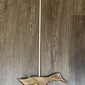 May include: Wooden toy duck on wheels with a yellow tail and a wooden stick handle. The duck is brown with white eyes.