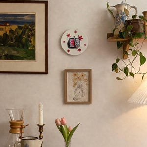 A decorative wall plate featuring a central illustration of a black and red toaster with red star accents. The plate is displayed on a wall alongside a framed landscape painting, a framed floral sketch, and a wooden shelf with a plant.