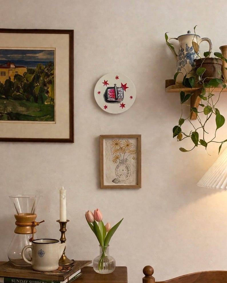 A decorative wall plate featuring a central illustration of a black and red toaster with red star accents. The plate is displayed on a wall alongside a framed landscape painting, a framed floral sketch, and a wooden shelf with a plant.