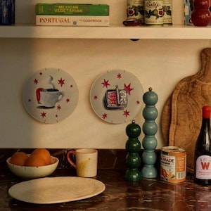 Two decorative wall plates featuring hand-painted illustrations of a steaming coffee cup with legs and a tin of sardines, both decorated with red stars. The plates are displayed on a wall above a kitchen counter with various items.
