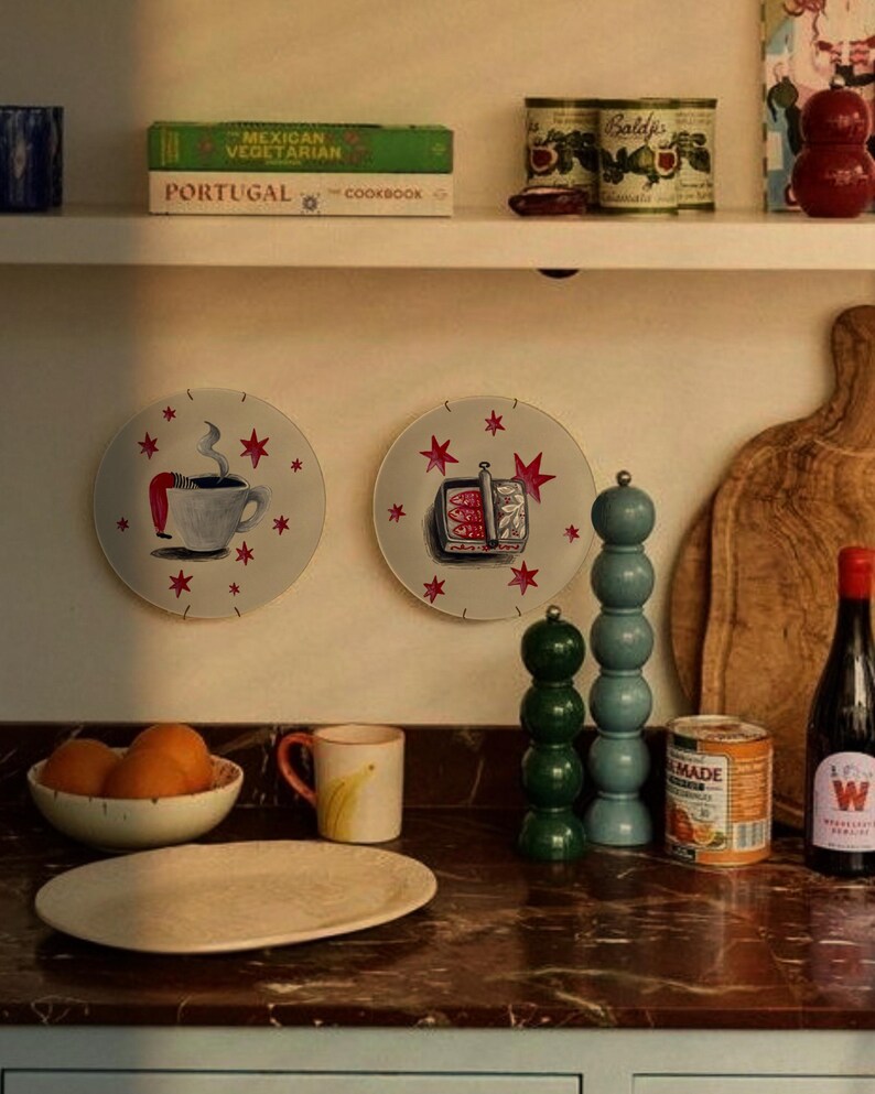 Two decorative wall plates featuring hand-painted illustrations of a steaming coffee cup with legs and a tin of sardines, both decorated with red stars. The plates are displayed on a wall above a kitchen counter with various items.