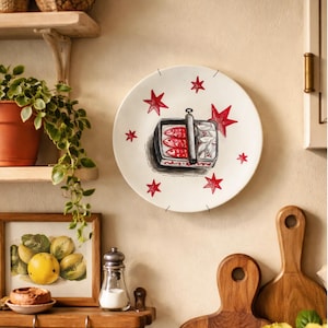 A round white ceramic decorative plate featuring a hand-painted illustration of an open tin of sardines in the center, surrounded by several red stars. The sardine tin is depicted with a silver lid and three red fish inside.