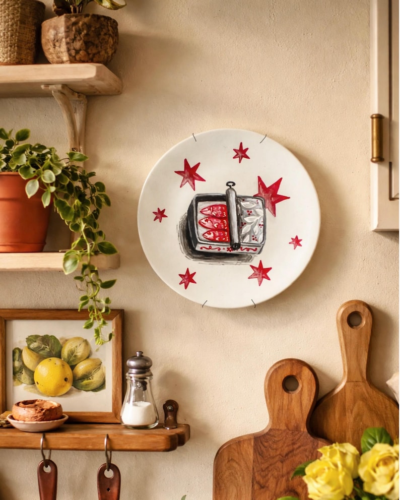 A round white ceramic decorative plate featuring a hand-painted illustration of an open tin of sardines in the center, surrounded by several red stars. The sardine tin is depicted with a silver lid and three red fish inside.