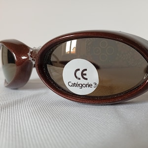 May include: Oval-shaped sunglasses with a dark brown, glittery frame and dark tinted lenses. A circular white sticker is applied to the right lens, featuring the text CE and Catégorie 3 in black, indicating the level of ultraviolet protection.