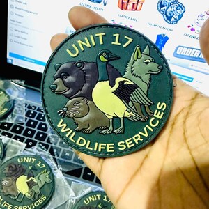 May include: A circular patch with the text "UNIT 17 WILDLIFE SERVICES." The patch features illustrations of a bear, a goose, a wolf, and a beaver in green, brown, and yellow. The patch is held in a hand.