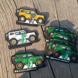 May include: Patches showing a cartoon dinosaur beside a yellow, white, and green SUV. The patches have the text "@GODZILLABRONCO" at the bottom. The patches are on a wooden surface.