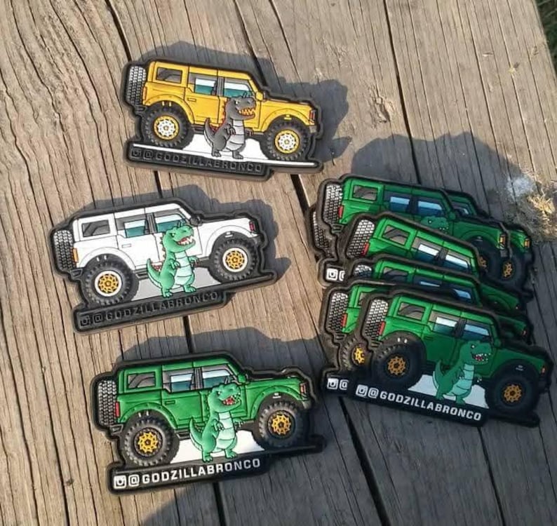 May include: Patches showing a cartoon dinosaur beside a yellow, white, and green SUV. The patches have the text "@GODZILLABRONCO" at the bottom. The patches are on a wooden surface.