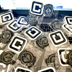 May include: A selection of black and white patches, some featuring a "C" design and others with a circular emblem reading "DEHCHO GUARDIAN". The patches are in clear plastic packaging, spread across a laptop keyboard.