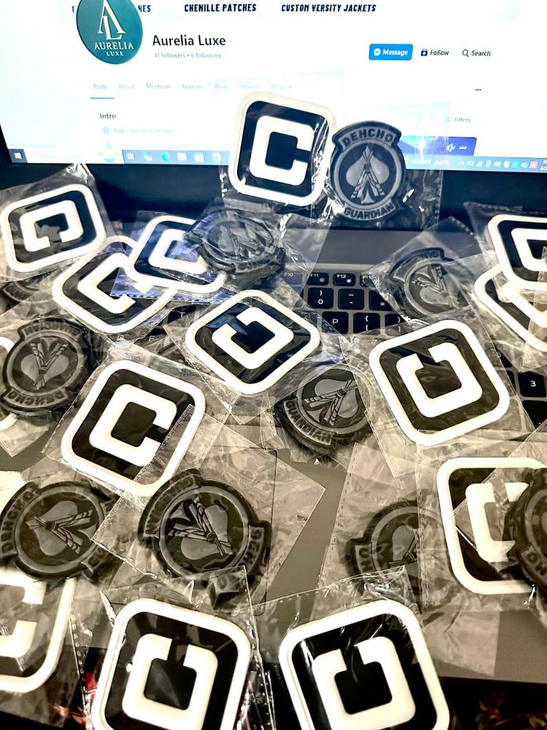 May include: A selection of black and white patches, some featuring a "C" design and others with a circular emblem reading "DEHCHO GUARDIAN". The patches are in clear plastic packaging, spread across a laptop keyboard.