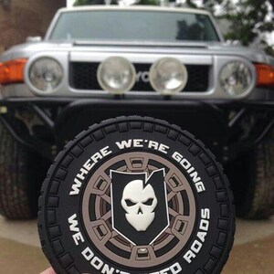 May include: A black, circular patch with the words "WHERE WE'RE GOING WE DON'T NEED ROADS" around a central emblem. The emblem shows a skull inside a shield. A silver SUV is visible in the background.