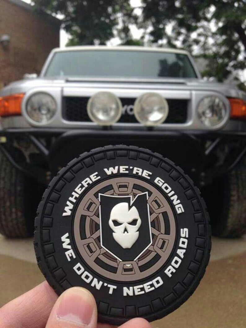 May include: A black, circular patch with the words "WHERE WE'RE GOING WE DON'T NEED ROADS" around a central emblem. The emblem shows a skull inside a shield. A silver SUV is visible in the background.