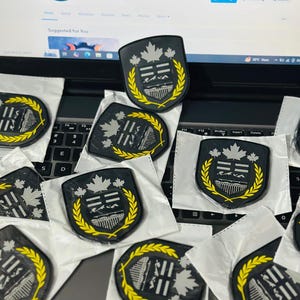 May include: Black and grey shield-shaped patches with a yellow laurel wreath border. The patches feature a silver maple leaf design. The patches are on a laptop keyboard.
