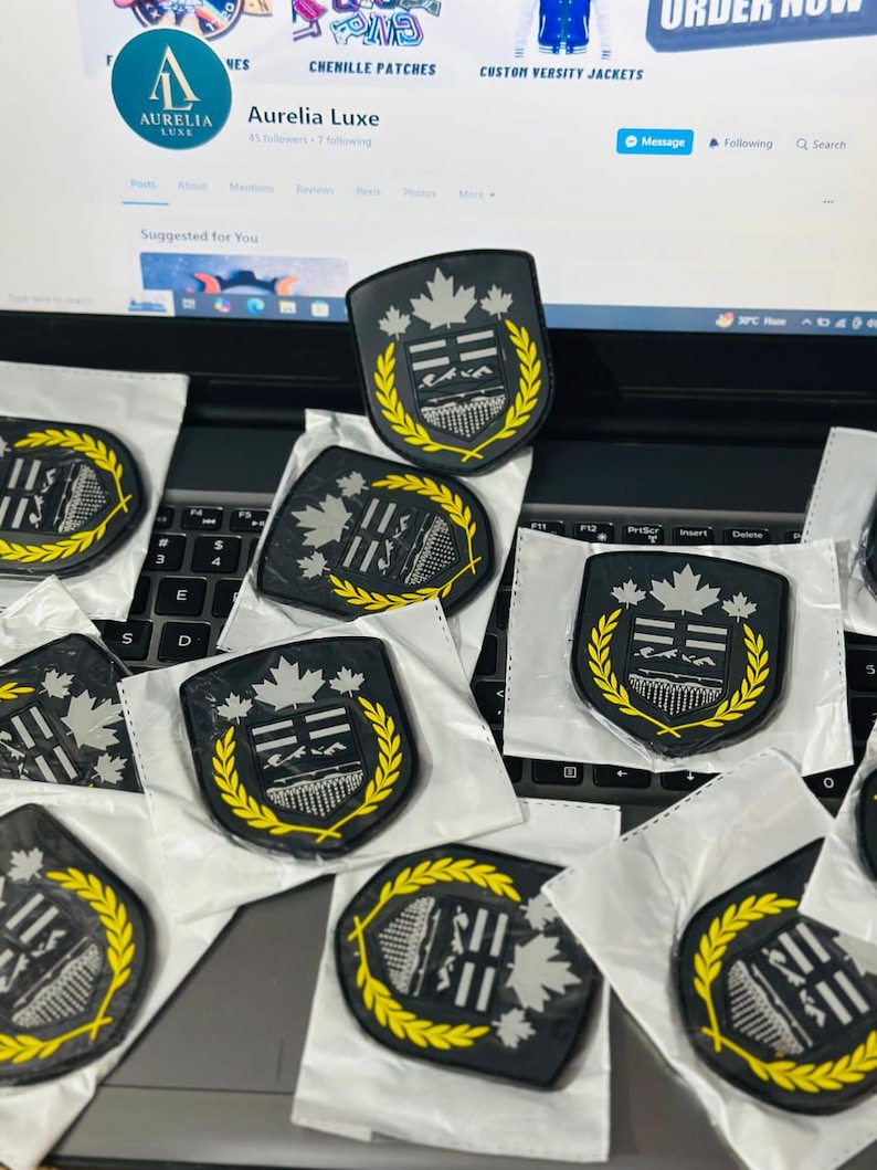 May include: Black and grey shield-shaped patches with a yellow laurel wreath border. The patches feature a silver maple leaf design. The patches are on a laptop keyboard.