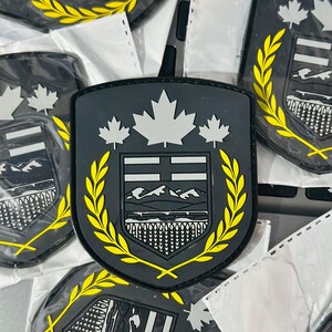 May include: Several black, shield-shaped patches are visible. Each patch has a white maple leaf design, a crest with a mountain and water scene, and a yellow laurel wreath border. The patches are in clear plastic packaging.