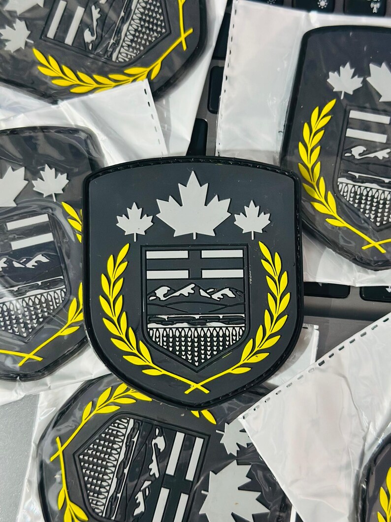May include: Several black, shield-shaped patches are visible. Each patch has a white maple leaf design, a crest with a mountain and water scene, and a yellow laurel wreath border. The patches are in clear plastic packaging.