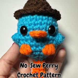 May include: A crocheted blue and orange Perry the Platypus amigurumi toy wearing a brown hat. The toy is holding its hands in front of it and has a small orange beak. The text "No-Sew Perry Crochet Pattern" is written below the toy.