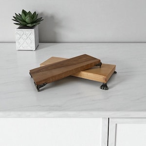 以下が含まれることがあります： 濃い茶色と薄い茶色の2つの木製サービングボードが表示されています。濃い茶色のボードが上にあり、明るい色のボードとわずかに重なっています。どちらのボードにも装飾的な金属製の足が付いています。背景には、多肉植物が入った白い正方形のプランターがあります。