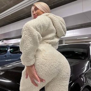 Könnte beinhalten: Cremefarbener, flauschiger Kapuzen-Overall. Das Outfit hat ein weiches, strukturiertes Aussehen. Das Model trägt den Overall in einer Parkgarage.