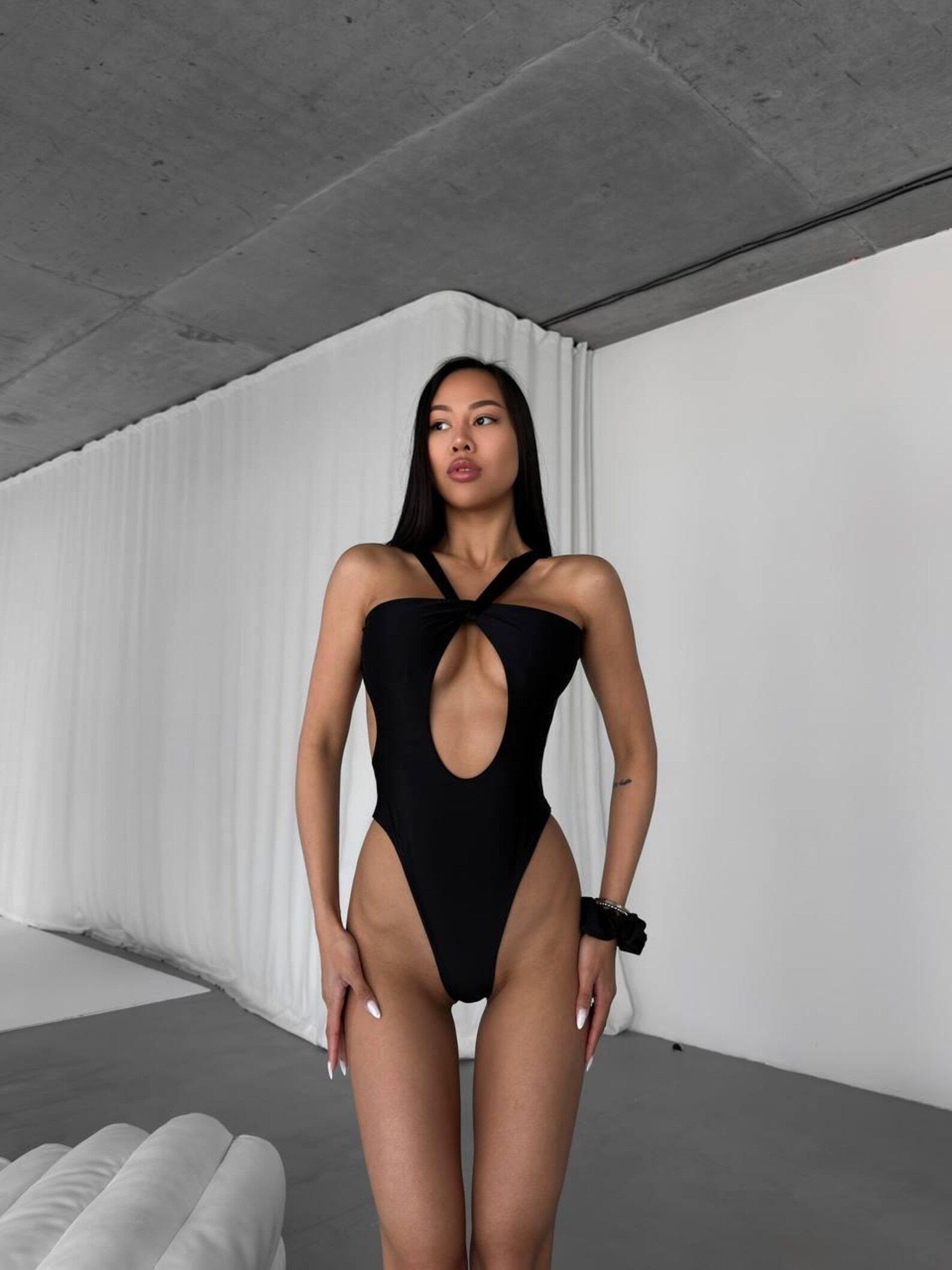 Woman wearing a black cutout monokini with a bold keyhole front and halter-style crossover straps, standing in a modern studio space.