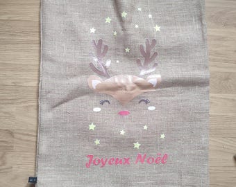 May include: A beige burlap Christmas gift bag with a reindeer face design, including antlers, eyes, and a pink nose. The bag has the text "Joyeux Noël" in pink and is decorated with yellow stars. The bag is tied at the top with a string.