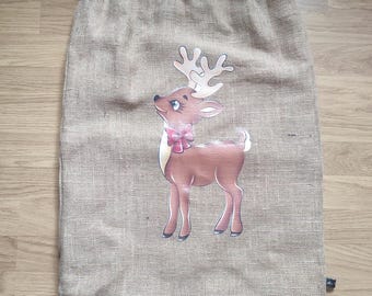 May include: A tan burlap Christmas gift bag with a drawstring closure. The bag features a cartoon reindeer illustration with a pink bow. The reindeer has large antlers and is standing on a white background.