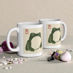 May include: Two white ceramic mugs with a green frog illustration on a beige background. The frog has a large, open mouth and is surrounded by a red border. The mugs are on a light-colored surface with decorative items.