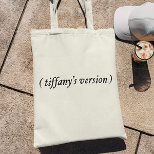 Könnte beinhalten: Cremefarbene Tragetasche mit dem Text "(tiffany's version)" in schwarzer Schrift. Die Tasche hat passende Griffe. Eine weiße Baseballkappe und ein Glas Eiskaffee sind ebenfalls zu sehen, die auf einer strukturierten Oberfläche liegen.