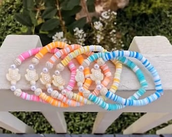 May include: A set of colourful bracelets with a layered design. Each bracelet features a white turtle charm and white beads. The bracelets are made of colourful, striped, and patterned beads.