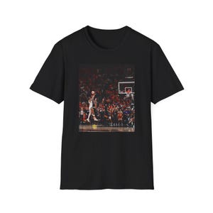 Puede incluir: Camiseta negra con una imagen de un partido de baloncesto. El gráfico muestra a un jugador saltando para lanzar el balón, con una multitud en el fondo. La camiseta tiene cuello redondo.