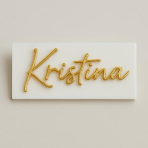 May include: A white rectangular name tag with the name "Kristina" in gold cursive lettering. The gold lettering is raised, creating a 3D effect. The name tag is simple and elegant.