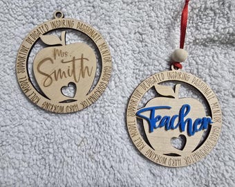 Può includere: Due ornamenti in legno a forma di mela. Uno presenta "Mrs. Smith" e un ritaglio a forma di cuore. L'altro dice "Teacher" in blu. Entrambi hanno un nastro rosso per appenderli e includono parole come "Dedicato" e "Ispiratore".