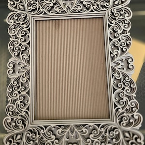 May include: Ornate, rectangular picture frame with a cream-colored, laser-cut design. The frame features intricate scrollwork and floral patterns around the edges, surrounding a tan, vertically-lined backing. The frame is designed to hold a photograph or artwork.