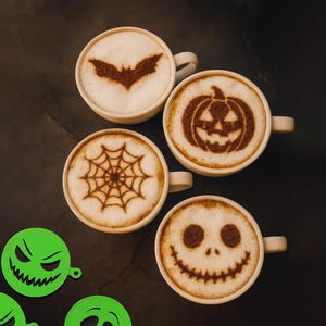 Peut inclure: Quatre tasses de café avec du latte art sur le thème d'Halloween. Les motifs comprennent une chauve-souris, une citrouille, une toile d'araignée et un visage souriant. Des pochoirs verts avec des visages effrayants sont au premier plan.