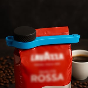Peut inclure: Un clip bleu avec un bouchon noir ferme un sac de café rouge. Une tasse de café blanche est en arrière-plan. Des grains de café sont éparpillés autour de la base du sac. Le clip est pour la conservation.
