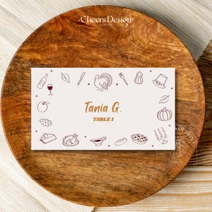 May include: A Thanksgiving-themed place card with the name "Tania G." and "Table 1" in gold lettering. The card is decorated with line drawings of Thanksgiving icons and sits on a wooden charger.
