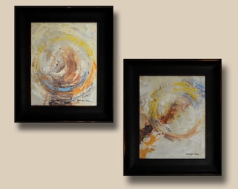 May include: Two framed abstract paintings with a swirling, circular design in shades of yellow, orange, brown, and blue. The paintings are set against a light beige wall.