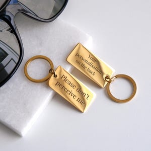 May include: Two gold-colored metal keychains with engraved text. The keychain on the left says "Please don't perceive me". The keychain on the right says "Your personalisation on the back".