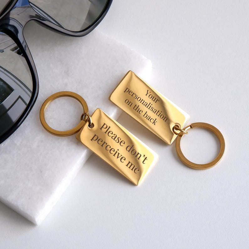 May include: Two gold-colored metal keychains with engraved text. The keychain on the left says "Please don't perceive me". The keychain on the right says "Your personalisation on the back".