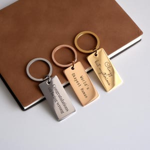 May include: Three silver, gold, and rose gold keychains with rectangular tags. The tags have engraved text: "Clingy but mysterious", "World's Okayest Human", and "Congratulations on being wrong".