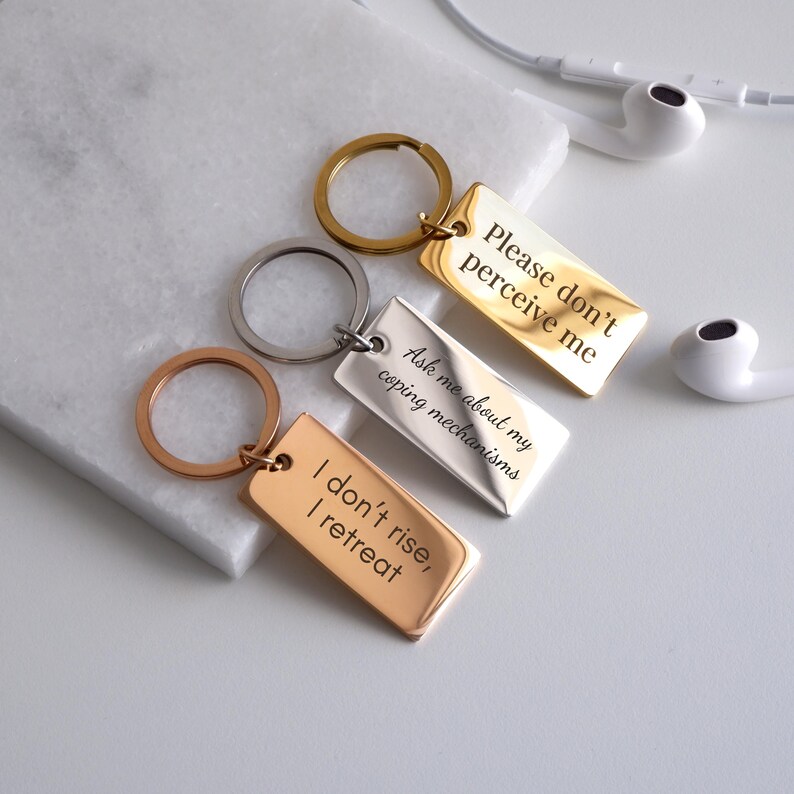 May include: Three gold, silver, and rose gold keychains with engraved sayings. The gold keychain says "Please don't perceive me". The silver keychain says "Ask me about my coping mechanisms". The rose gold keychain says "I don't rise, I retreat".