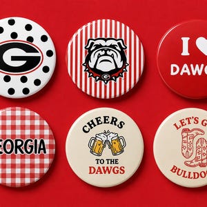 May include: Six circular pinback buttons featuring various designs: a black G on white polka dots, a bulldog on red stripes, a red button with I heart DAWGS, a red gingham button with GEORGIA, a cream button with beer mugs and CHEERS TO THE DAWGS, and cowboy boots with LET'S GO BULLDOGS.